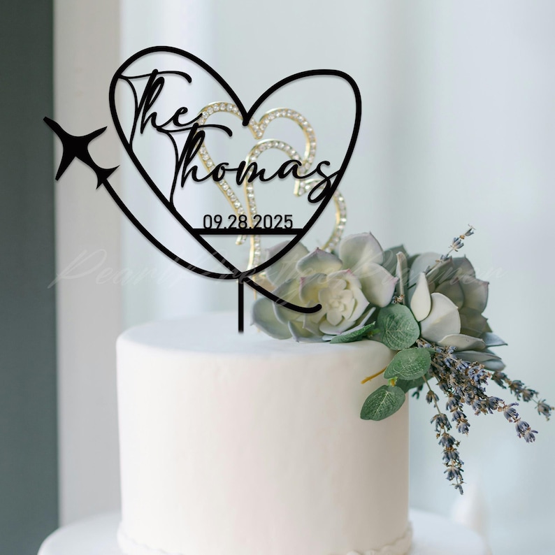 May include: A black cake topper featuring a heart outline, a silhouette of an airplane, and the text "The Thomas" above the date "09.28.2025". The topper is positioned on a white cake decorated with a cluster of green succulents and lavender.