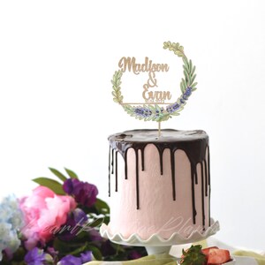 May include: A pink cake with chocolate ganache dripping down the sides, topped with a wooden cake topper. The topper features the names "Madison & Evan" and the date "10.04.2023" within a floral wreath design.