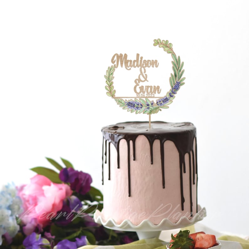 May include: A pink cake with chocolate ganache dripping down the sides, topped with a wooden cake topper. The topper features the names "Madison & Evan" and the date "10.04.2023" within a floral wreath design.