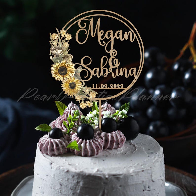 Puede incluir: Una tarta de boda adornada con un adorno de pastel de madera. El adorno presenta los nombres "Megan & Sabrina" en escritura elegante, rodeados de un dise&ntilde;o floral con girasoles. Tambi&eacute;n se incluye la fecha "11.09.2022".