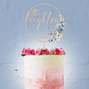 May include: A wooden cake topper with the text "The Keighley" and the date "10.26.2022" within a circular frame. The frame is decorated with blue and cream-colored floral accents. The cake is topped with pink roses.