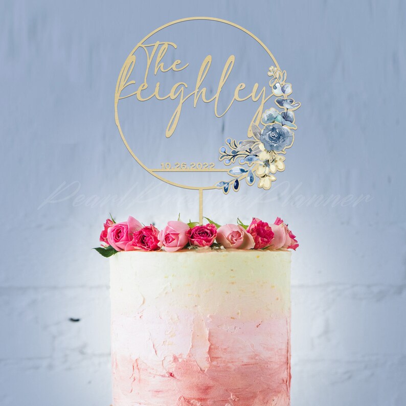 May include: A wooden cake topper with the text "The Keighley" and the date "10.26.2022" within a circular frame. The frame is decorated with blue and cream-colored floral accents. The cake is topped with pink roses.