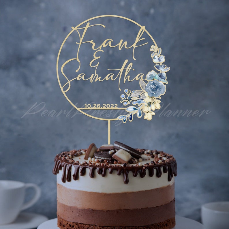 May include: A gold-colored cake topper with the names "Frank & Samantha" and the date "10.26.2022" is displayed on top of a chocolate cake. The topper is circular with floral accents in blue and cream. The cake has three layers and is topped with chocolate.