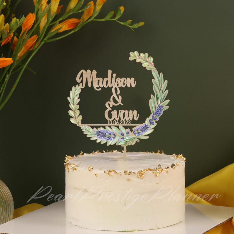 May include: A white frosted cake with gold leaf accents, topped with a wooden cake topper. The topper features the names "Madison & Evan" and the date "10.06.2023" within a floral wreath design. The cake is a wedding or celebration cake.
