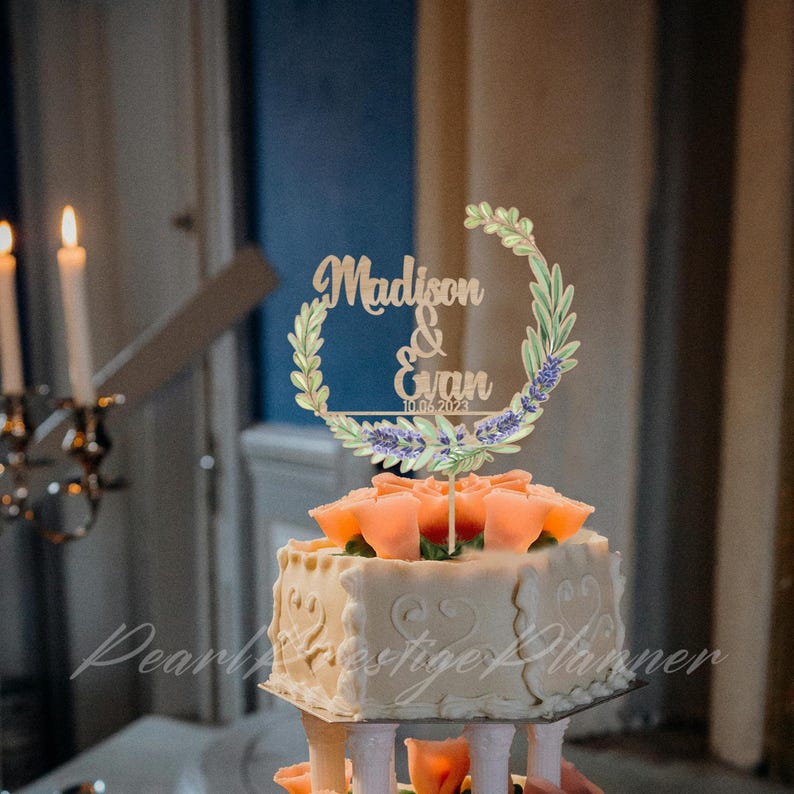 May include: A multi-tiered wedding cake with peach-colored roses and a decorative topper. The topper features the names "Madison & Evan" and the date "10.06.2023" within a floral wreath. The cake has a white frosting with decorative patterns.