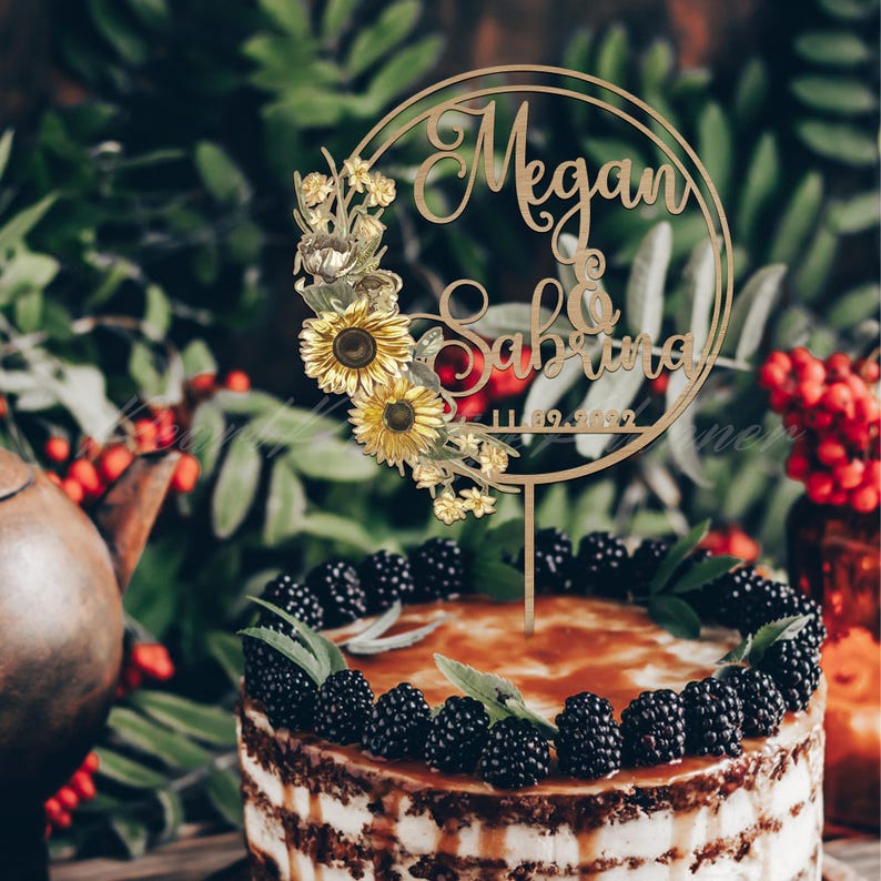 Puede incluir: Un adorno de pastel de madera con los nombres "Megan & Sabrina" y la fecha "11.09.2022" en un dise&ntilde;o circular, adornado con girasoles y detalles florales. El adorno est&aacute; colocado sobre un pastel decorado con moras.