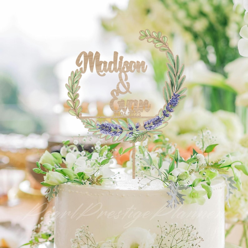 May include: A wedding cake with a decorative topper that reads "Madison & Evan" in a script font. The topper is a wooden design with a floral wreath of lavender and greenery. The cake is white and decorated with white flowers.