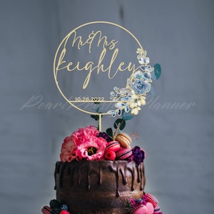 May include: A gold-colored cake topper with the words "Mr & Mrs Keighley" and the date "10.26.2022" inside a circular frame. The frame is decorated with blue and white floral accents. The cake is topped with pink flowers, macarons, and berries.