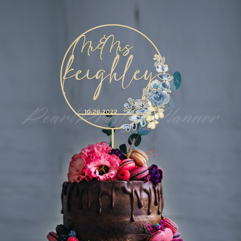 May include: A gold-colored cake topper with the words "Mr & Mrs Keighley" and the date "10.26.2022" inside a circular frame. The frame is decorated with blue and white floral accents. The cake is topped with pink flowers, macarons, and berries.