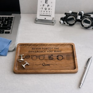 May include: A rectangular wooden tray with the engraved text "NEVER FORGET THE DIFFERENCE YOU MAKE" and the name "Quinn". The tray features engraved images of eyeglasses, an eye, and other optometry-related symbols. Two silver cufflinks are on the tray.