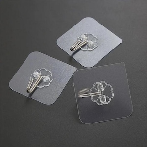 May include: Three clear adhesive wall hooks, each featuring a square transparent backing plate and a silver-toned metal hook attached to a flower-shaped base. The hooks are designed for mounting on flat surfaces, providing a functional solution.