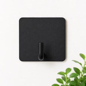 May include: A matte black square wall hook featuring a flat, rounded-corner base plate and a single, centred cylindrical hook extending outwards. The design is simple and functional, providing a clean look for hanging various household items.