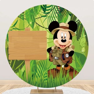 May include: A round backdrop featuring a cartoon image of Mickey Mouse dressed as an explorer in a jungle setting. Mickey is wearing a brown hat, a green shirt, and brown pants. He is holding a compass in his hand. There is a wooden sign in the background.