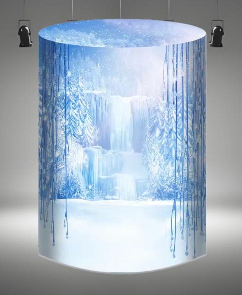 May include: A cylindrical backdrop with a winter scene featuring a waterfall and snow-covered trees. The background is a light blue with white snowflakes and a light pink sky. The bottom of the backdrop is white and appears to be snow.