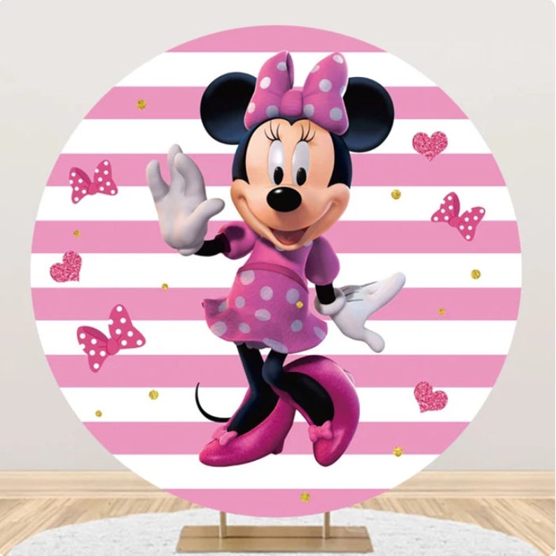 May include: A pink and white striped circular backdrop featuring a cartoon image of Minnie Mouse. Minnie Mouse is wearing a pink polka dot dress, pink shoes, and a pink bow with white polka dots. The backdrop is decorated with pink hearts and gold glitter.