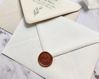 Peut inclure: Deux enveloppes couleur crème en papier texturé. Une enveloppe présente un texte imprimé en noir indiquant kindly respond to Aaron and Maria, 42 Willow Lane, Ashcombe, West Mercia, XM1 2ZZ et une illustration florale. L'autre a un sceau en cire cuivré.