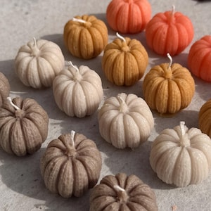 May include: A collection of small, pumpkin-shaped candles in various colours including shades of orange, yellow, beige, brown, and cream. Each candle features a ribbed texture and a single white wick centred at the top of the pumpkin shape.
