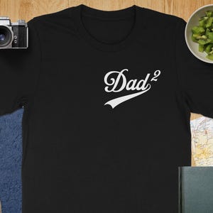 May include: Black t-shirt with the white text "Dad²" in a script font with a baseball-style underline. The shirt is displayed on a wooden surface with a camera, lens, and a bowl of kiwi.