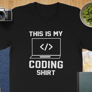 May include: Black t-shirt with the text "THIS IS MY CODING SHIRT" in white. The design includes a laptop graphic with code symbols. A camera, lens, and a bowl of kiwi are on the wooden surface.