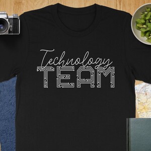 May include: Black t-shirt with the words "Technology TEAM" in white script and circuit board style lettering. The shirt is displayed on a wooden surface with a camera, lens, and a bowl of kiwi.