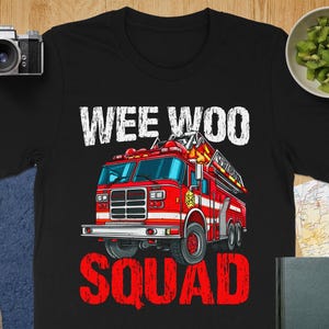 May include: Black t-shirt featuring a red fire truck illustration with the words "WEE WOO SQUAD" in white and red. The fire truck has a ladder and a star emblem. The shirt is on a wooden surface with a camera and a bowl of kiwi.