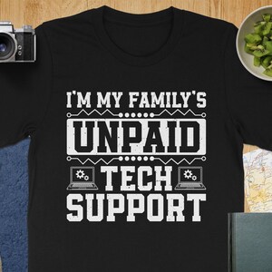 May include: Black t-shirt with the white text "I'M MY FAMILY'S UNPAID TECH SUPPORT." The design includes laptop graphics. The shirt is a crew neck and appears to be made of cotton or a similar material. The shirt is displayed on a wooden surface.