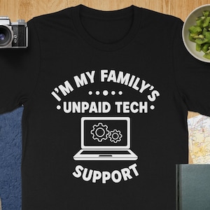 May include: Black t-shirt with the white text "I'M MY FAMILY'S •UNPAID TECH• SUPPORT" and a laptop graphic with gears. The shirt is a crew neck and is displayed on a wooden surface.