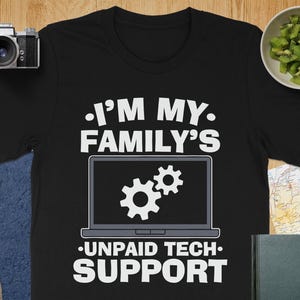 Puede incluir: Camiseta negra con el texto "I'M MY FAMILY'S UNPAID TECH SUPPORT" en blanco. El diseño presenta un portátil con dos iconos de engranajes en la pantalla. La camiseta está sobre una superficie de madera con una cámara, una lente y un cuenco de kiwi.