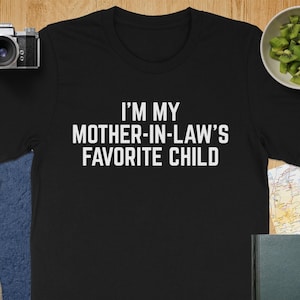 May include: Black t-shirt with the white text "I'M MY MOTHER-IN-LAW'S FAVORITE CHILD." The shirt is on a wooden surface with a camera, lens, and a bowl of kiwi. A map and a book are also visible.
