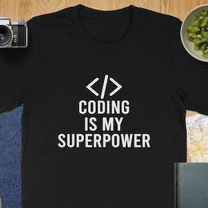 May include: Black t-shirt with the text "CODING IS MY SUPERPOWER" in white. The shirt features a coding symbol above the text. The image also includes a camera, lens, and a bowl of kiwi.