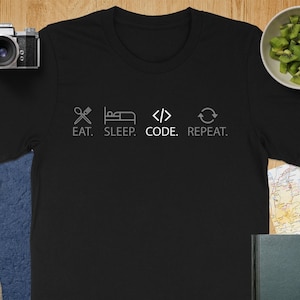 May include: Black crew neck t-shirt with the text "EAT. SLEEP. CODE. REPEAT." in a light gray color. The design includes icons representing a fork and knife, a bed, code brackets, and a circular arrow. The shirt is on a wooden surface.