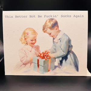 May include: Greeting card with a vintage illustration of two children opening a gift. The gift is wrapped in blue paper with a red bow. The text on the card reads: "This Better Not Be Fuckin' Socks Again."