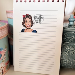 May include: A spiral-bound notepad with cream-colored lined paper and pink plastic binding rings. The top section features a retro-style illustration of a smiling person with a red hair bow and a speech bubble that reads, WELL, SLAP MY KNEE AND CALL ME SALLY!