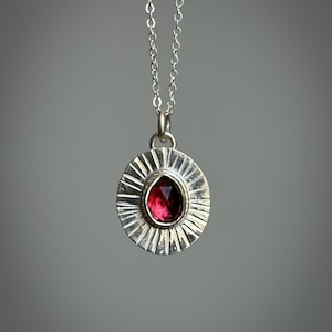 Pu&ograve; includere: Una collana con pendente in argento con una gemma rosso scuro a forma di goccia. Il pendente &egrave; incastonato in una cornice circolare con un design a raggi di sole. La catena della collana &egrave; delicata e argentata.