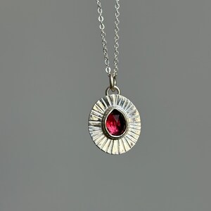 Pu&ograve; includere: Collana con pendente in argento con un pendente rotondo con un design a raggi di sole. Il pendente contiene una gemma rosso scuro a forma di goccia. La catena della collana &egrave; sottile e argentata.