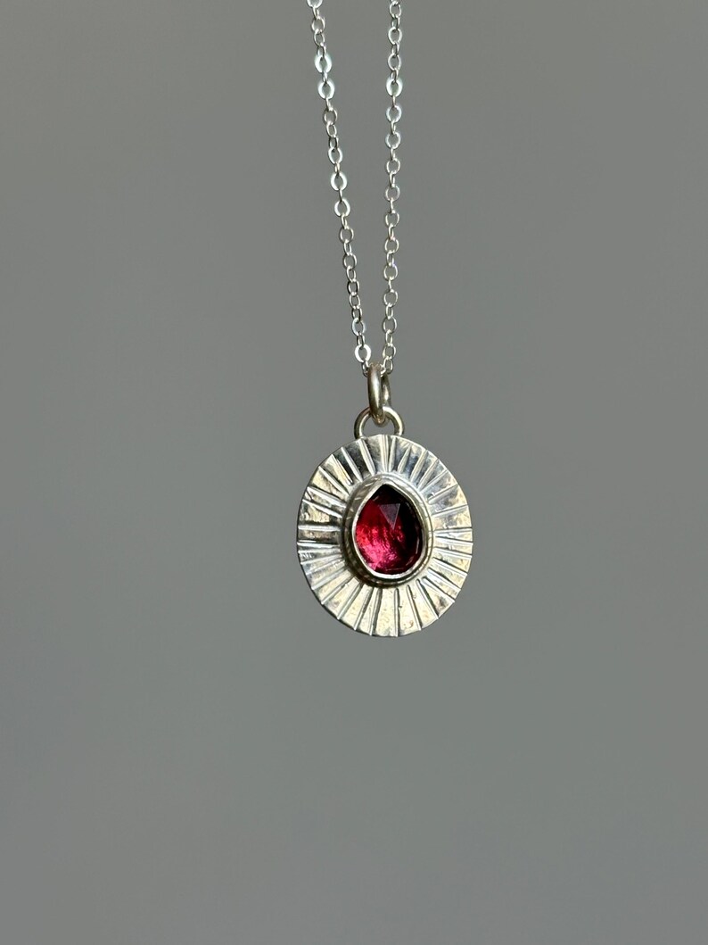 Pu&ograve; includere: Collana con pendente in argento con un pendente rotondo con un design a raggi di sole. Il pendente contiene una gemma rosso scuro a forma di goccia. La catena della collana &egrave; sottile e argentata.