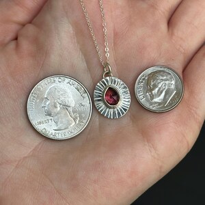 Pu&ograve; includere: Una collana d'argento con un ciondolo con una gemma rossa a forma di goccia. Il ciondolo &egrave; circondato da un design a forma di raggio di sole. Due monete sono accanto al ciondolo per il confronto delle dimensioni.
