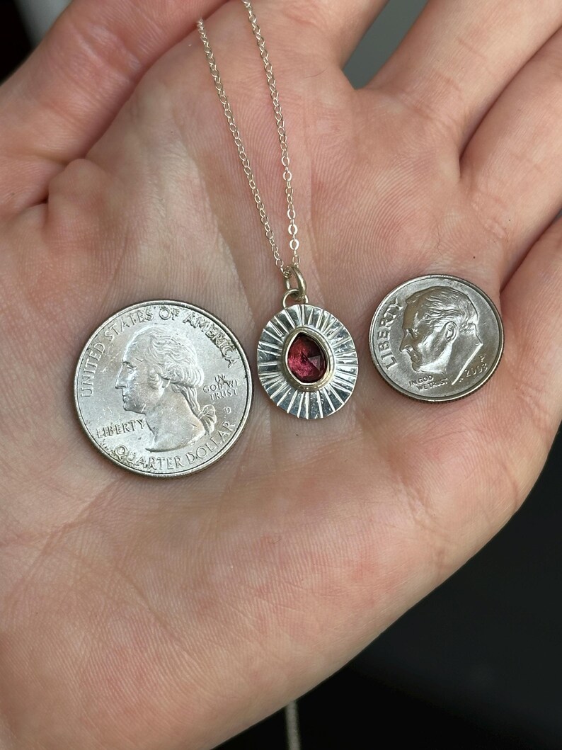 Pu&ograve; includere: Una collana d'argento con un ciondolo con una gemma rossa a forma di goccia. Il ciondolo &egrave; circondato da un design a forma di raggio di sole. Due monete sono accanto al ciondolo per il confronto delle dimensioni.