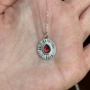 Pu&ograve; includere: Una collana d'argento con un pendente rotondo con una pietra preziosa rossa. Il pendente ha un design a raggiera attorno alla gemma. La collana &egrave; mostrata su una mano.