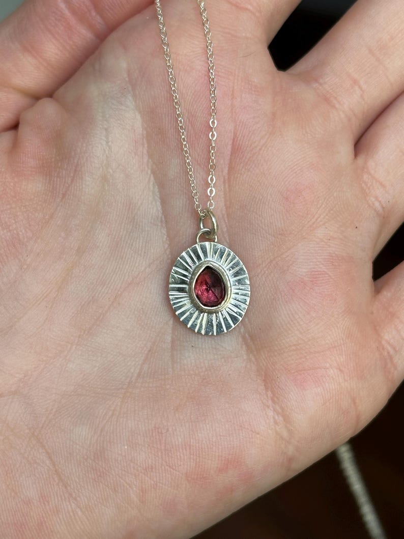 Pu&ograve; includere: Una collana d'argento con un pendente rotondo con una pietra preziosa rossa. Il pendente ha un design a raggiera attorno alla gemma. La collana &egrave; mostrata su una mano.