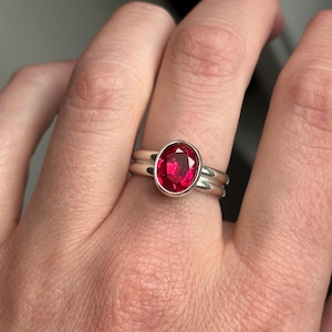 May include: A silver ring featuring a large, oval-cut, vibrant red gemstone. The ring has a simple, elegant design with a smooth, polished band. The gemstone is set in a bezel setting, enhancing its color and brilliance. A second band is visible.