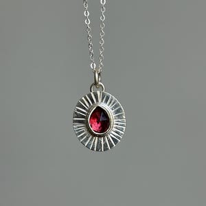 Pu&ograve; includere: Una collana con pendente in argento con una pietra preziosa rosso scuro a forma di goccia incastonata in un design a raggiera. Il pendente &egrave; appeso a una delicata catena d'argento, creando un gioiello elegante.