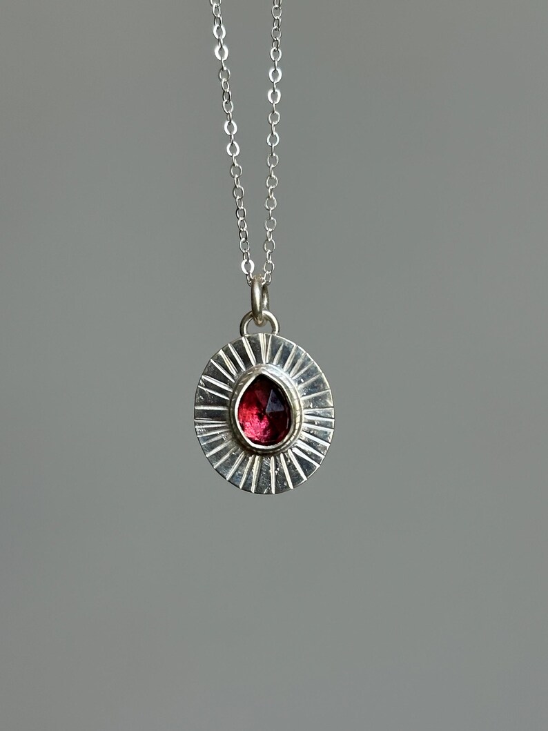 Pu&ograve; includere: Una collana con pendente in argento con una pietra preziosa rosso scuro a forma di goccia incastonata in un design a raggiera. Il pendente &egrave; appeso a una delicata catena d'argento, creando un gioiello elegante.