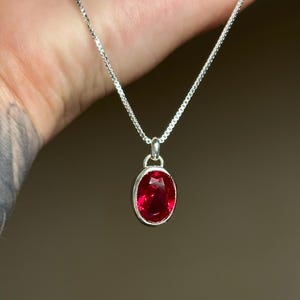 May include: A silver necklace with a bright red oval pendant. The pendant is set in a silver bezel and hangs from a delicate silver chain. The gemstone has a faceted surface.