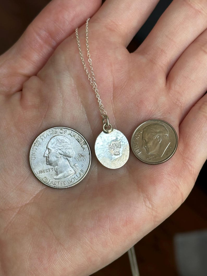 Pu&ograve; includere: Una collana d'argento con un piccolo ciondolo rotondo. Il ciondolo ha le iniziali "AJM" e "925". Una moneta da un quarto di dollaro e una da dieci centesimi sono accanto al ciondolo per il confronto delle dimensioni.
