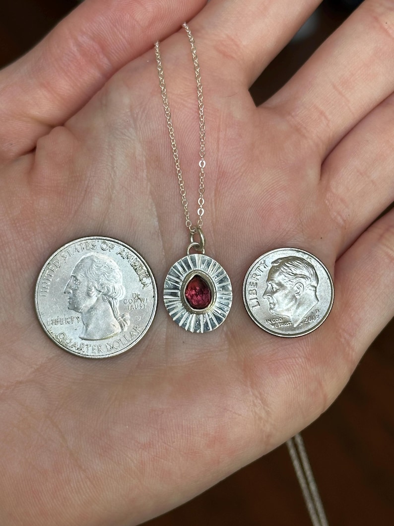 Pu&ograve; includere: Una collana con pendente in argento con una gemma rossa al centro. Il pendente &egrave; rotondo con un bordo strutturato. La collana &egrave; mostrata con un quarto di dollaro e una monetina da dieci centesimi per il confronto delle dimensioni.