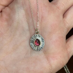 Pu&ograve; includere: Collana con pendente in argento con una gemma rosso scuro a forma di goccia. Il pendente ha un design a raggiera attorno alla pietra e la catena &egrave; delicata. Un gioiello.