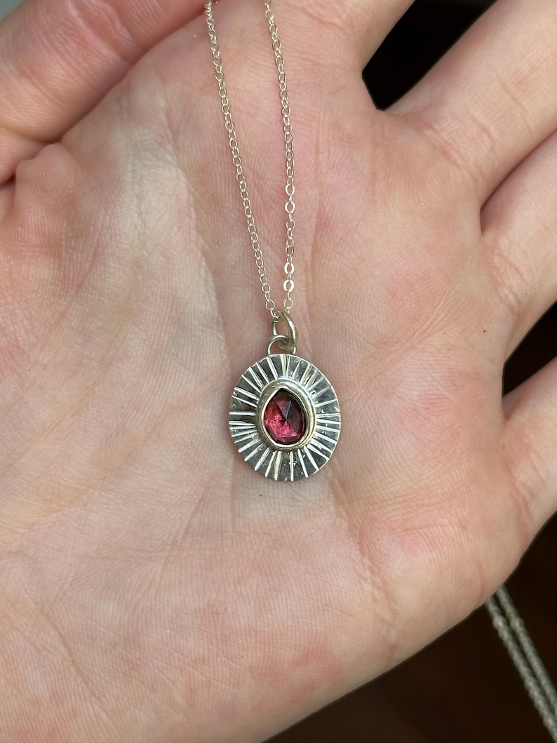 Pu&ograve; includere: Collana con pendente in argento con una gemma rosso scuro a forma di goccia. Il pendente ha un design a raggiera attorno alla pietra e la catena &egrave; delicata. Un gioiello.