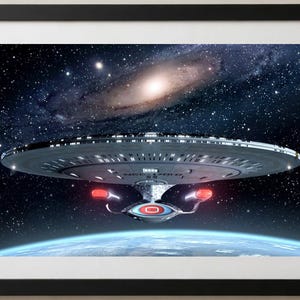 May include: A framed print featuring the Starship Enterprise NCC-1701-D in deep space. The saucer-shaped vessel is positioned above a blue planet with a bright galaxy and dense star field in the background. The ship displays red engine glows.