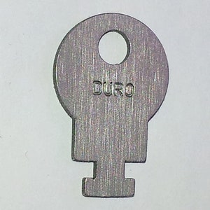 May include: A silver key with a round head and a T-shaped bottom. The key has the word "DURO" engraved on the head.